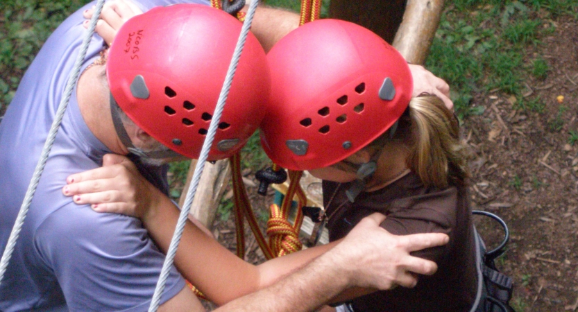 Two people wearing safety gear are secured by ropes as they make their way through a high ropes course. They are facing each other, bracing themselves on each other's shoulders and are pressing their helmets together.